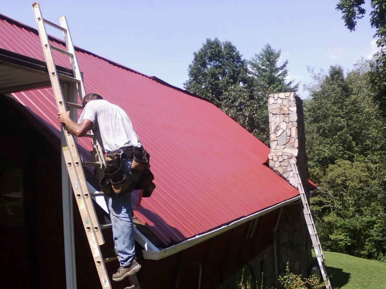 Roofer on a ladder beside a red metal roof and stone chimney — Soffit & Fascia Repair by Roofing Brothers in Poteet